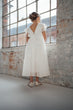 A young girl stands with her back to the camera wearing a white tulle dress with lace puff sleeves and a lace-up open back, in a rustic brick and glass room.