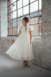 A young girl wearing a white lace and tulle first communion dress stands in a rustic industrial loft with exposed brick walls and large multi-pane windows.