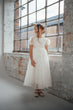 A young girl wearing a white tulle first birthday dress with short puff sleeves and a lace bodice, standing in a bright room with a large industrial window and brick walls.