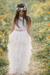 Flower girl in a white dress with a floral crown standing in a field of flowers.