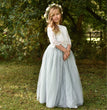 Young girl in a white lace top and light green tulle skirt standing outdoors with greenery in the background.