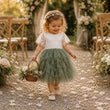 A young girl with curly blonde hair and a floral headband walks down a flower-paved aisle carrying a small wicker basket. She is wearing a white lace short-sleeve top and a tiered sage green tulle tutu skirt.