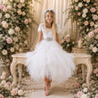 A young girl stands barefoot in a white tulle dress with a silver beaded waistband and floral crown, posed on a carved bench between large cream and pink floral arrangements.