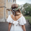 Child in a white dress with floral hairpiece standing in front of a historic building.