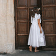 A young girl seen from behind wearing a white short-sleeve dress with a sheer tulle overskirt, standing barefoot in front of large dark wooden doors.