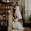 Two young girls in white sleeveless dress gowns sit and stand on stone steps inside a church or historic interior, with a wooden pulpit and stone walls in the background.