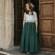 A young girl wearing a bohemian-style two-tone dress with a white lace bodice and a dark green full skirt, standing barefoot in a rustic doorway with wooden doors and a stone wall.