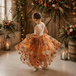 A young girl viewed from behind wearing a sleeveless white lace top and a voluminous orange tulle skirt with floral embroidery, twirling in a rustic barn decorated with lanterns and flowers.