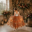 A young girl stands with her back to the camera, holding out a voluminous orange tulle dress with a floral print and a white lace bodice, in a rustic indoor setting decorated with flowers and lanterns.