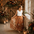 A young girl stands by a window in a rustic indoor setting, wearing a sleeveless white top and a long rust-orange tulle skirt with a floral print. The scene includes lanterns, candles, wooden doors, and large floral arrangements.