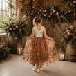 A young girl stands with her back to the camera in a rustic indoor setting, wearing a sleeveless white lace bodice and a full rust-orange tulle skirt with floral embroidery. She is surrounded by warm boho decor, including wooden barrels, lanterns with candles, and large floral arrangements.