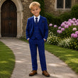 Young boy in a blue suit standing on a path with flowers and a building in the background