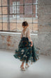 A young girl viewed from behind wearing a dark teal tulle flower girl dress with white daisy embroidery and a lace bodice, standing in a bright studio with brick walls and a large window.