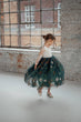 A young girl twirling in a white long-sleeve top and a voluminous dark green tulle skirt decorated with white and orange floral patterns, standing in a bright studio with brick walls and a large window.