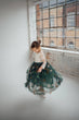 A young girl twirling in a white lace long-sleeve top and a voluminous dark green tulle skirt decorated with floral embroidery, standing in a bright room with a large industrial window.
