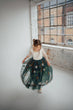 A young girl twirling in a voluminous dark green tulle dress with a cream lace bodice, standing in a bright studio with a large industrial window.