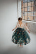 A young girl viewed from behind wearing a long-sleeved lace top and a full dark green tulle skirt decorated with white daisy-like flowers and orange floral accents, standing in a bright room with a large window.