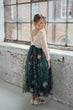A young girl wearing a long-sleeved lace top and a dark teal tulle skirt with orange and white floral embroidery, standing in a rustic room with a large window.
