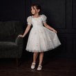 Young girl in a white floral dress standing in a room with dark walls and a green couch.