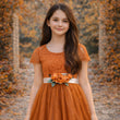 Young girl in an orange dress with a floral sash standing in an autumn setting.