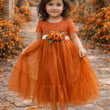 Young girl in an orange dress with a floral sash standing in a natural setting with autumn foliage.
