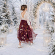 A woman stands in a snowy outdoor aisle wearing a sleeveless burgundy tulle maxi skirt with floral embroidery and a matching lace back bodice. The scene is decorated with white lanterns, snow-covered trees, and a glowing arched backdrop.