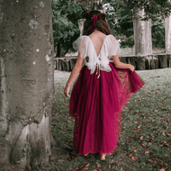A girl wearing a burgundy high low dress with a layered tulle skirt and off white quality lace upper bodice, with a lace tie and ruffle detail, standing next to a tree.