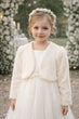 Young girl in a white dress with a fur shrug standing in front of floral decorations.