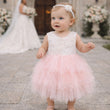 Baby in a pink and white dress standing outdoors with blurred wedding background