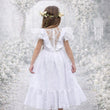 Young girl in a white dress with lace details standing against a textured white background.