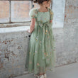 Young girl in a green floral dress standing against a brick wall.