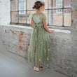 Young girl in a green floral dress standing by a window with a brick wall background