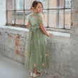 Young girl in a green floral dress standing in front of a brick wall with large windows.