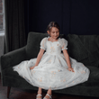 Young girl in a white dress with floral embroidery sitting on a dark green couch.