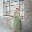 Young girl in a green floral dress standing in front of large windows with a brick wall background.