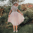 A woman stands outdoors in a garden wearing a short-sleeve light pink dress with a lace back bodice and a full tulle skirt, holding out the sides of the skirt to show its volume. She has a floral crown and is wearing white flats.
