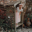 A young girl wearing a light pink tulle dress and a floral crown stands on a wooden garden bench inside a rustic wooden garden shed, surrounded by a garden with potted plants and autumn foliage.